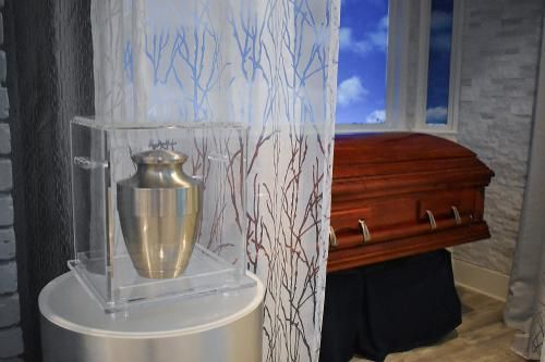 A metallic cremation urn in a clear case sits on a pedestal next to a wood-finish casket in a room with a window.
