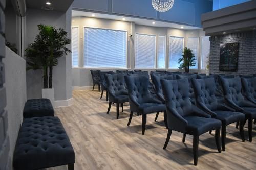 An elegant room with rows of blue tufted chairs, light wood flooring, floor-to-ceiling windows with blinds, and plants.
