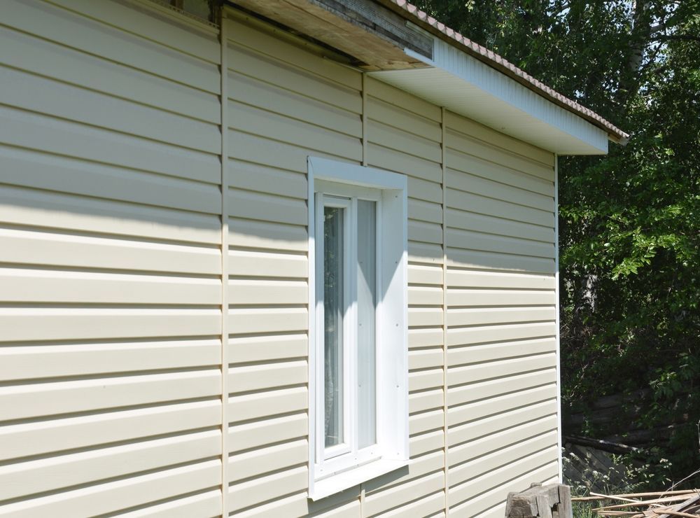 Side of a beige house with white siding and a white window in daylight
