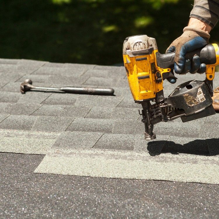 Worker using a yellow nail gun to install gray roof shingles on a sunny day