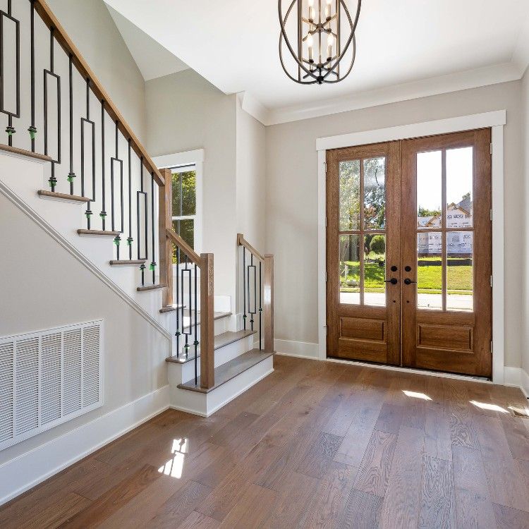 Bright entryway with hardwood floors, staircase, and double wooden front doors opening to a sunny yard