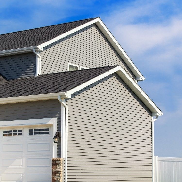 Suburban house exterior with gray siding, dark shingle roof, and white garage door under a blue sky
