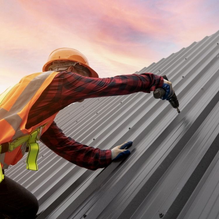 Worker in orange safety gear installing a corrugated metal roof with a drill.