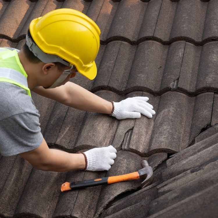 Worker in a yellow hard hat inspecting roof shingles with a hammer nearby