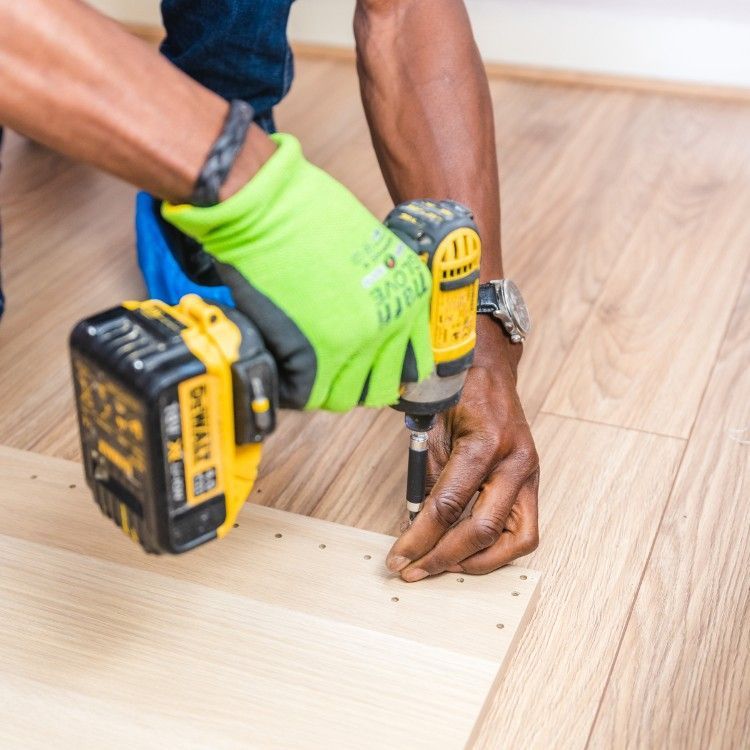 Worker using a drill to fasten a wooden floorboard, wearing green gloves and black shoes.