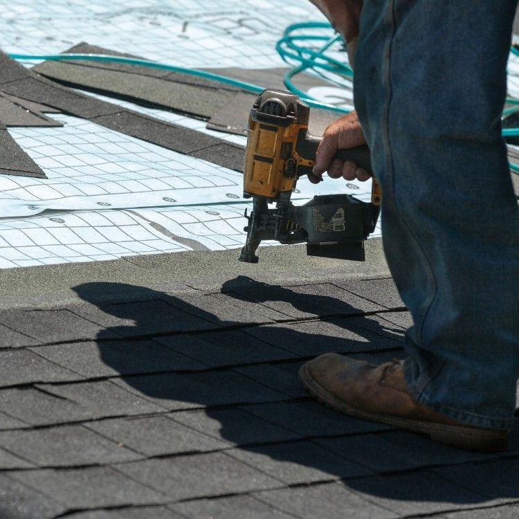 Worker using a nail gun to install shingles on a roof, with coiled hose and tools nearby