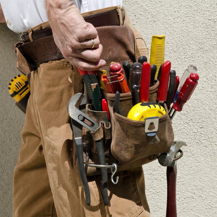 A carpenter’s tool belt with pliers, screwdrivers, tape measure, and a hand holding it against a wall.