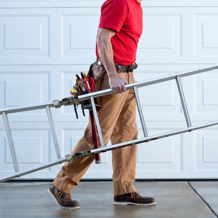 Worker carrying a metal ladder in a garage, wearing a red shirt and tan pants.