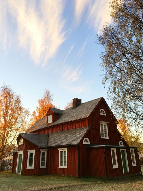 Red wooden cottage with white trim and a steep roof among autumn trees under a blue sky