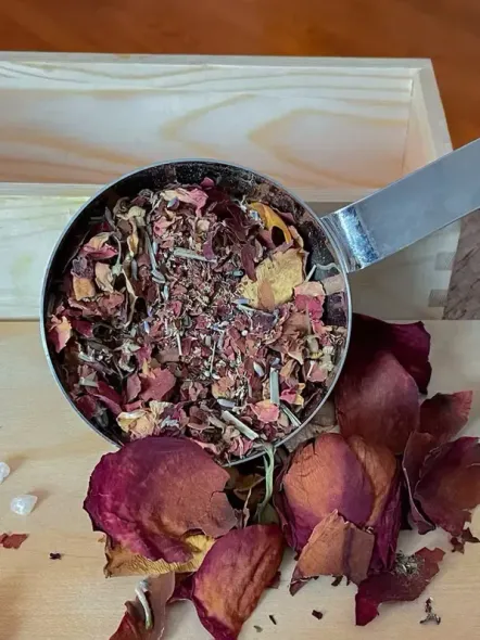 Metal scoop filled with dried rose petals and herbs, spilling onto a wooden surface alongside scattered petals.