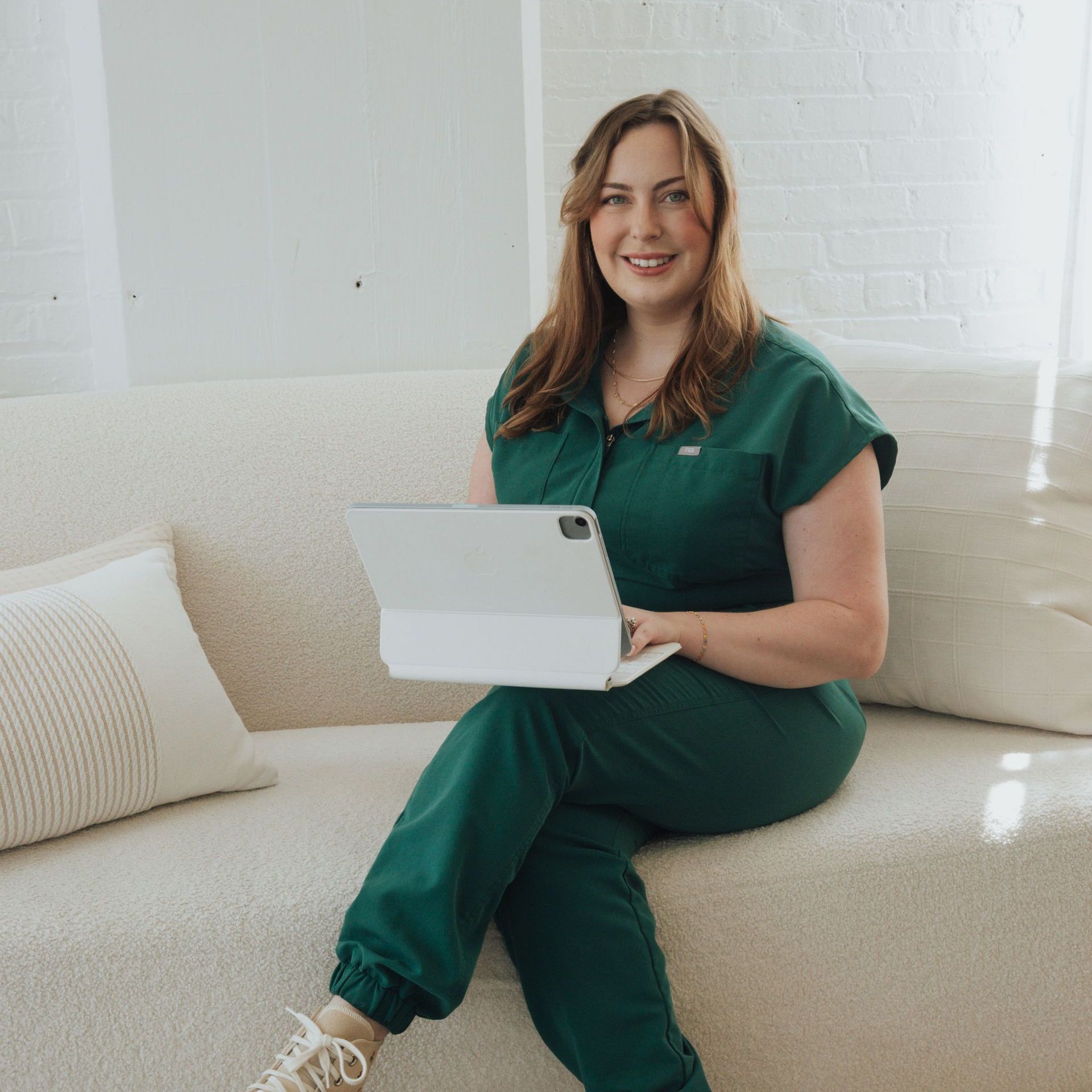 Woman in green jumpsuit smiles while working on a tablet, seated on a beige couch.