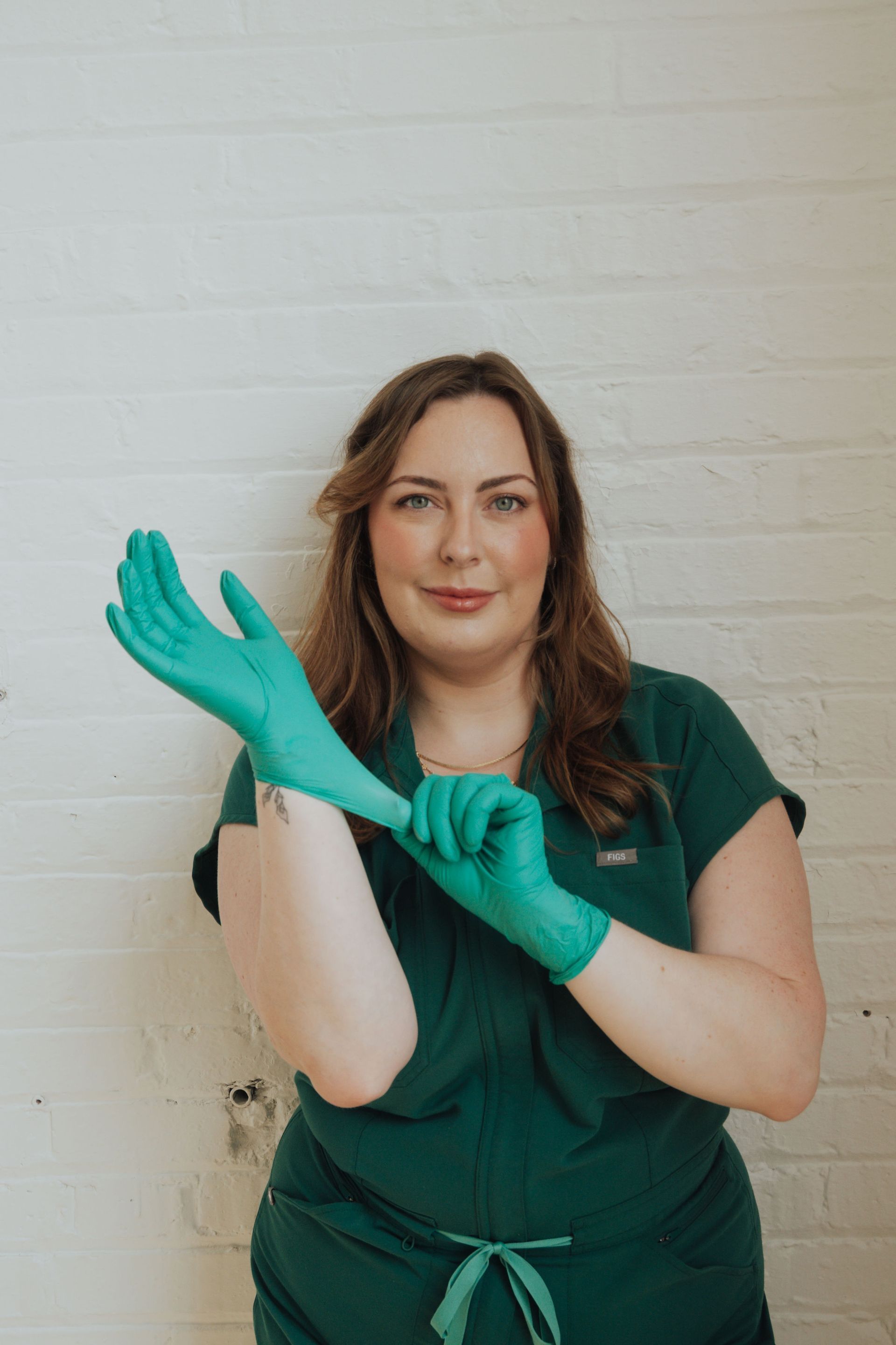 Woman in green scrubs putting on a teal glove against a white brick wall.