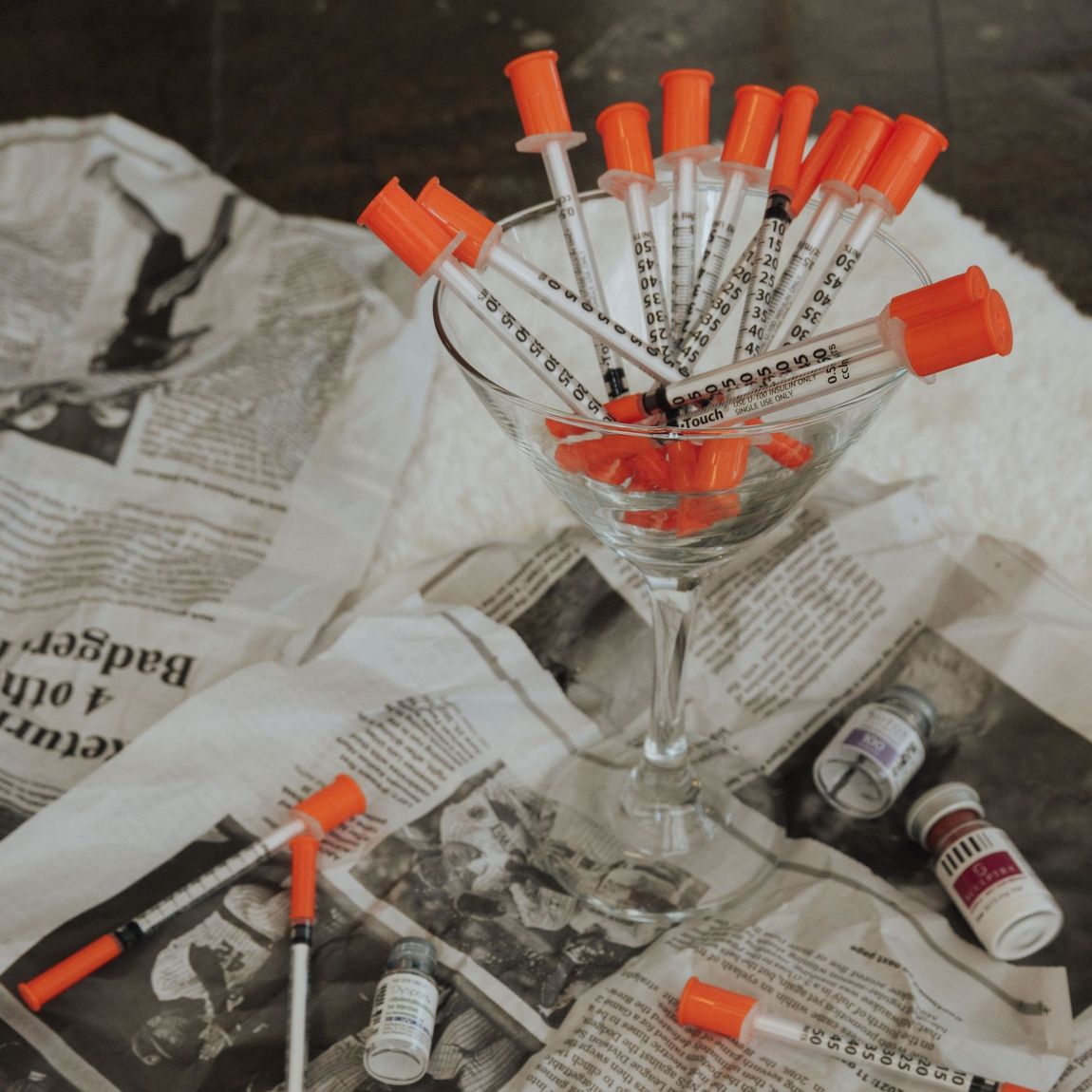 A martini glass filled with insulin syringes, spilled on a newspaper-covered surface with vials.