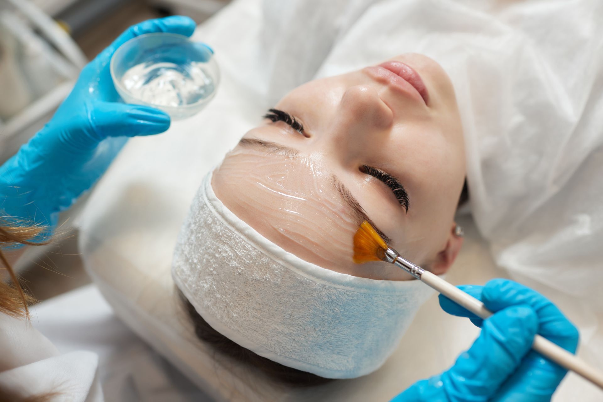 Person receiving facial peel, skin treatment with brush and gel, in a clinic, wearing gloves.