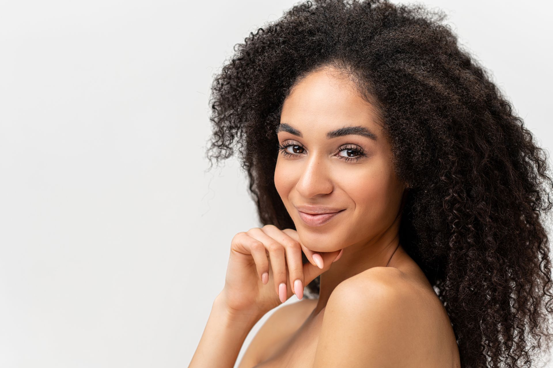 Woman with dark, curly hair smiles, hand on chin, against a white background.