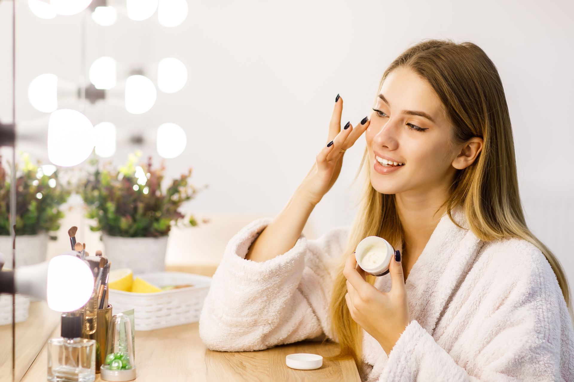 Woman in a white robe applying makeup at a vanity with lit bulbs and cosmetics.
