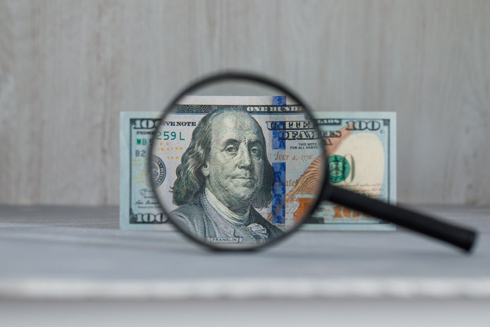 Magnifying glass over a $100 bill, Benjamin Franklin's portrait visible, set against a neutral backdrop.