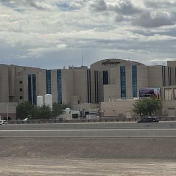 Large beige stadium with blue accents under cloudy sky, viewed from across a highway.