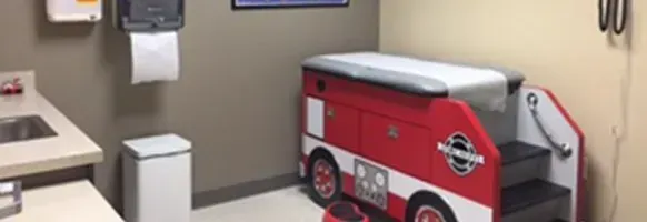 Red toy ambulance in a clinic exam room beside a sink and paper towel dispenser