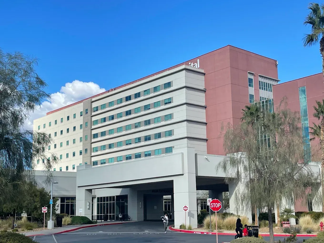 Hospital building with white and pink wings, entrance canopy, and a stop sign under a clear blue sky
