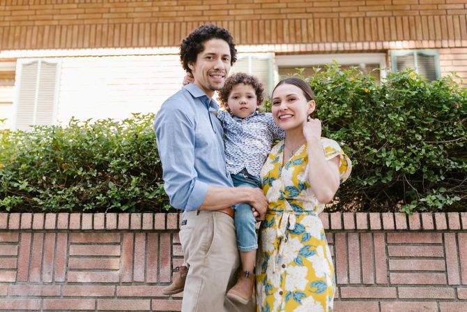 Smiling family of three standing by a brick wall and greenery, with a child held between them