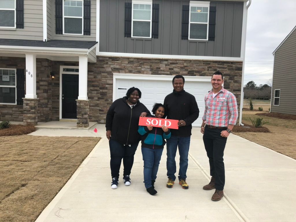Four people stand in front of a house holding a red “SOLD” sign on the driveway.