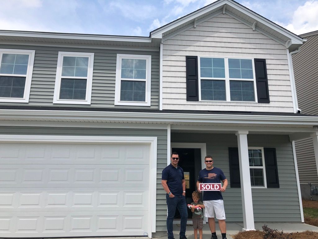 Two people standing in front of a gray house with a two-car garage, one holding a red and white sign.