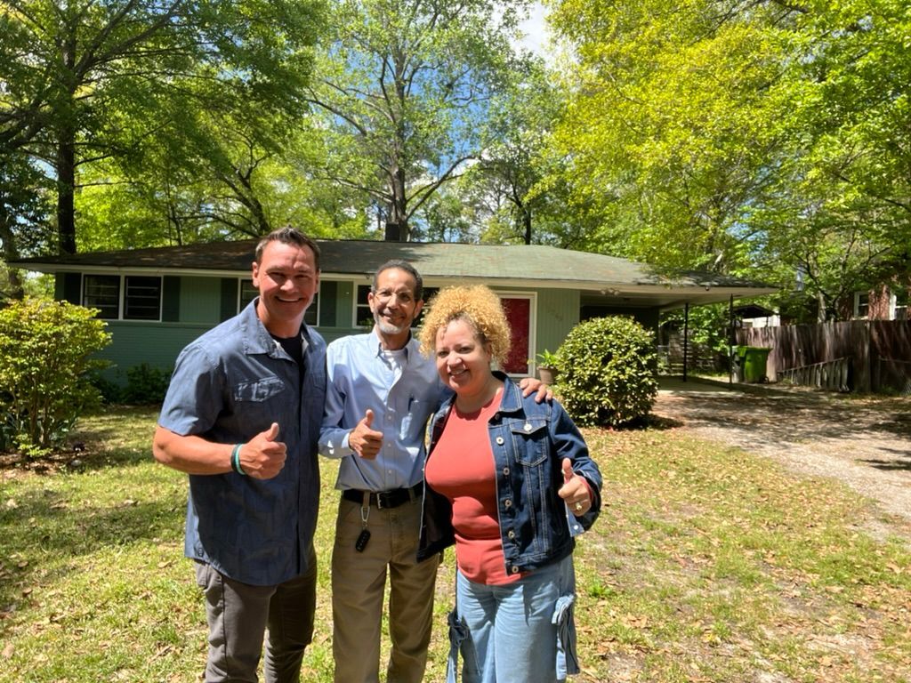 Three people smiling and giving thumbs-up in front of a house and trees on a sunny day