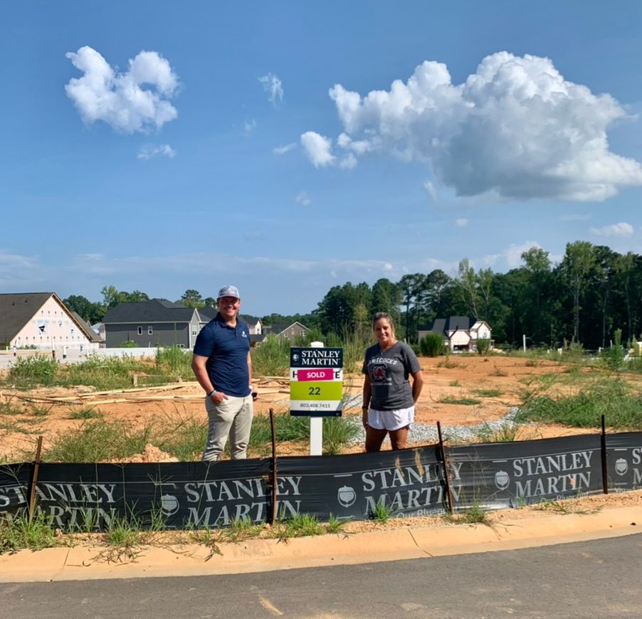 Two people stand at a bare lot with a real estate sign under a blue sky and clouds.