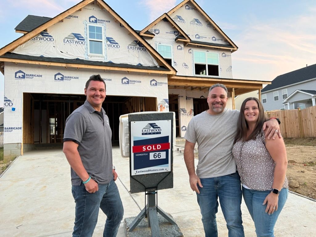 Three people standing in front of a sold house sign and a new home under construction.