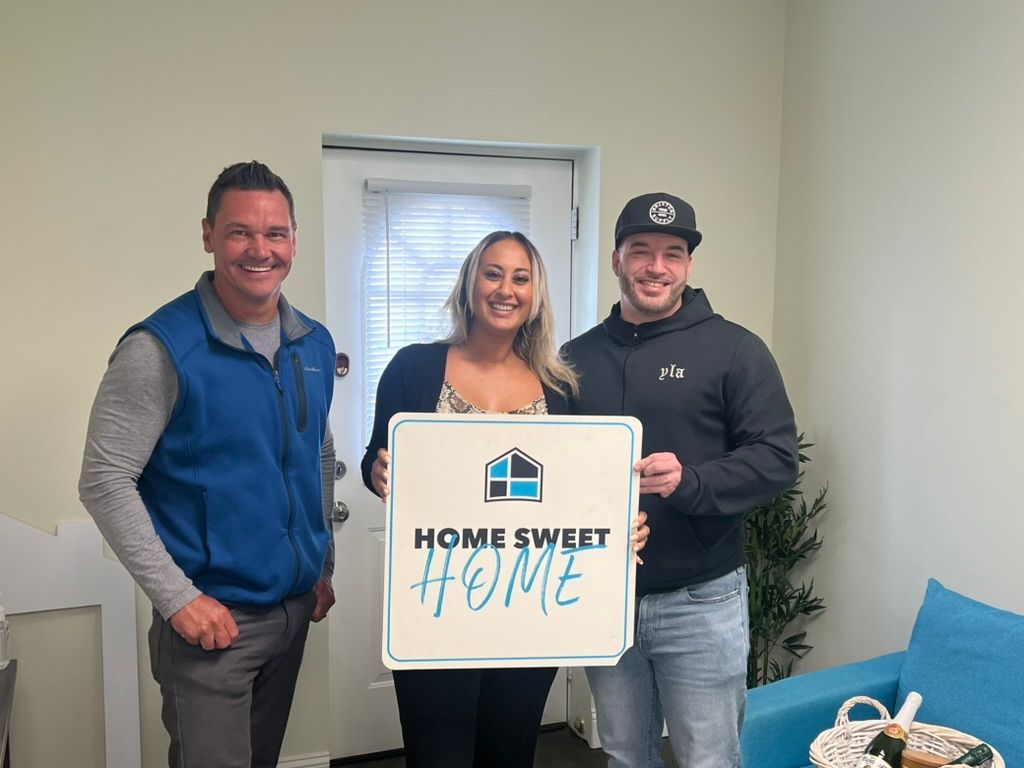 Three people in a room holding a “Home Sweet Home” sign, smiling beside a couch.
