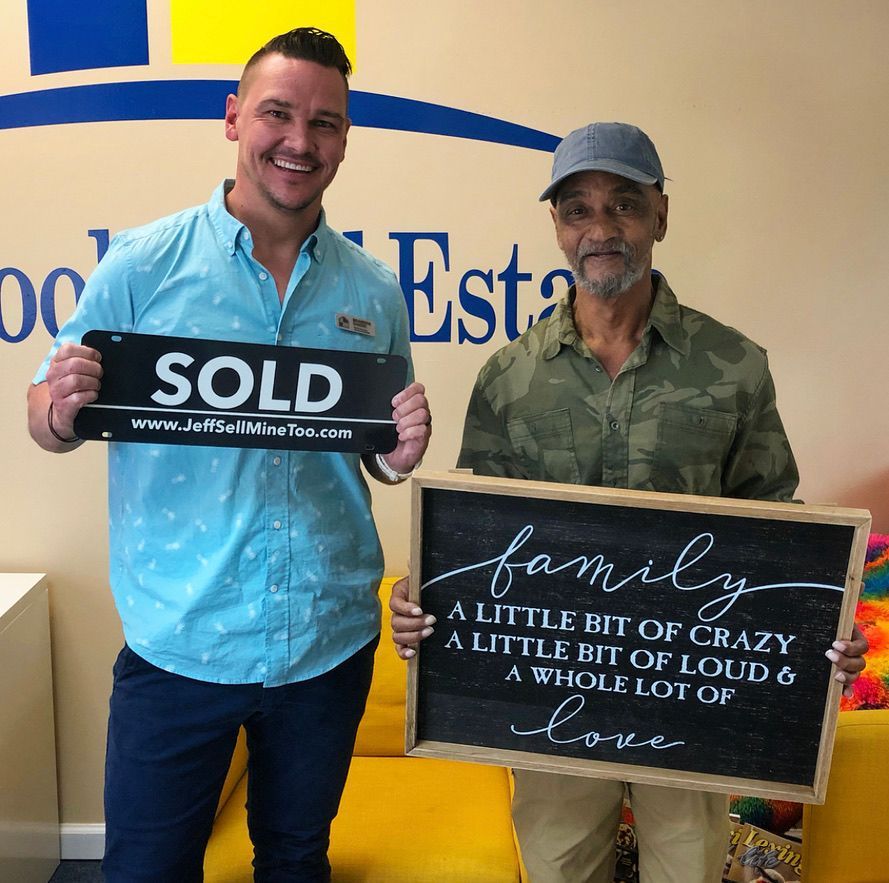Two men hold real estate signs: “SOLD” and “family...a little bit of crazy...” in an office.