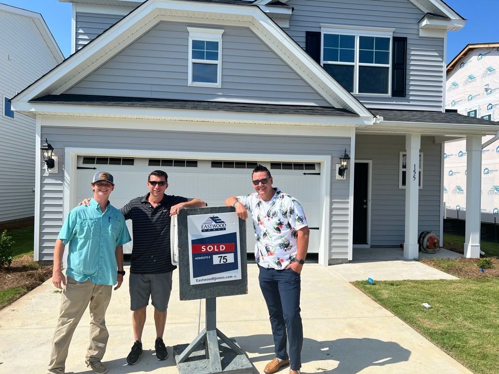 Three men standing beside a real estate sign in front of a gray house and garage.