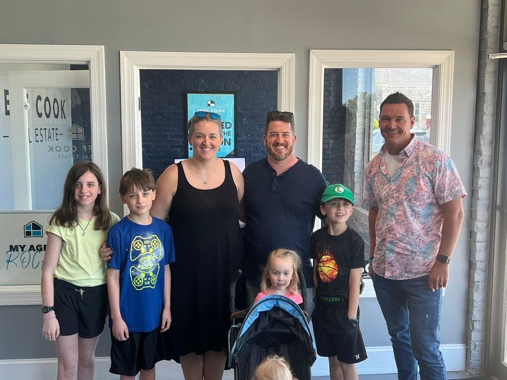 Group of six smiling people standing outside a storefront doorway, with one child in a stroller in front