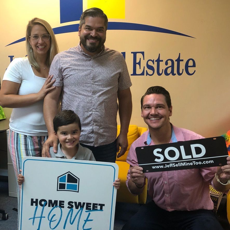 Family holding real estate signs in front of an estate office, with a smiling agent holding a SOLD sign.