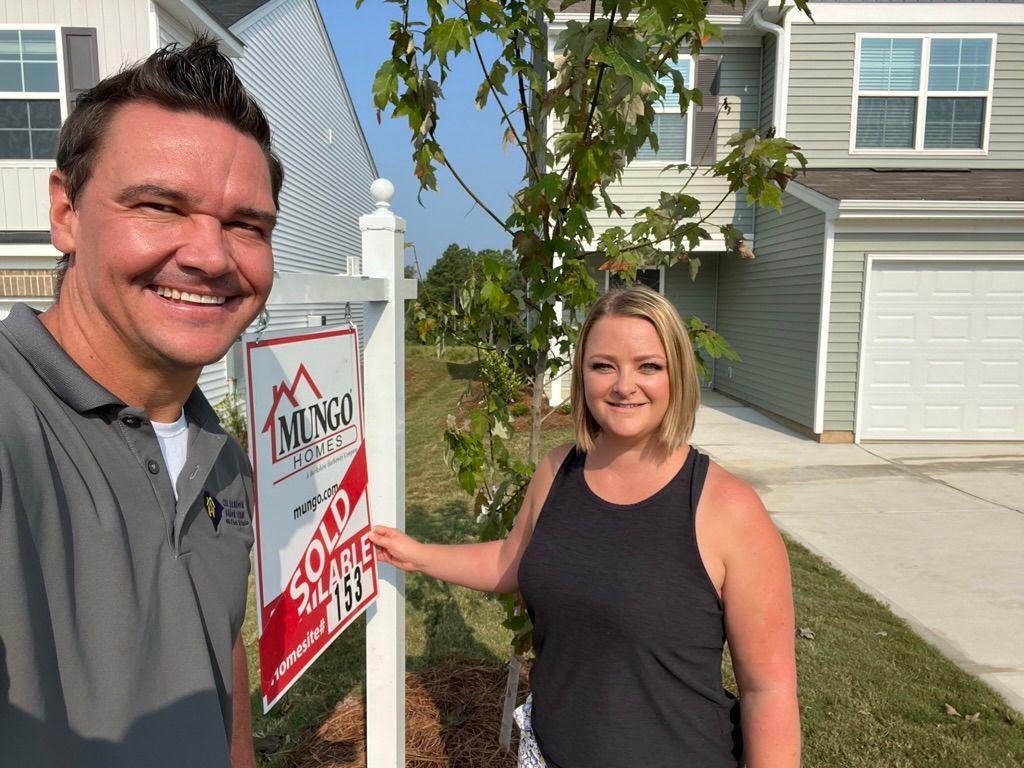 Two people smiling beside a real estate sign in a suburban neighborhood street