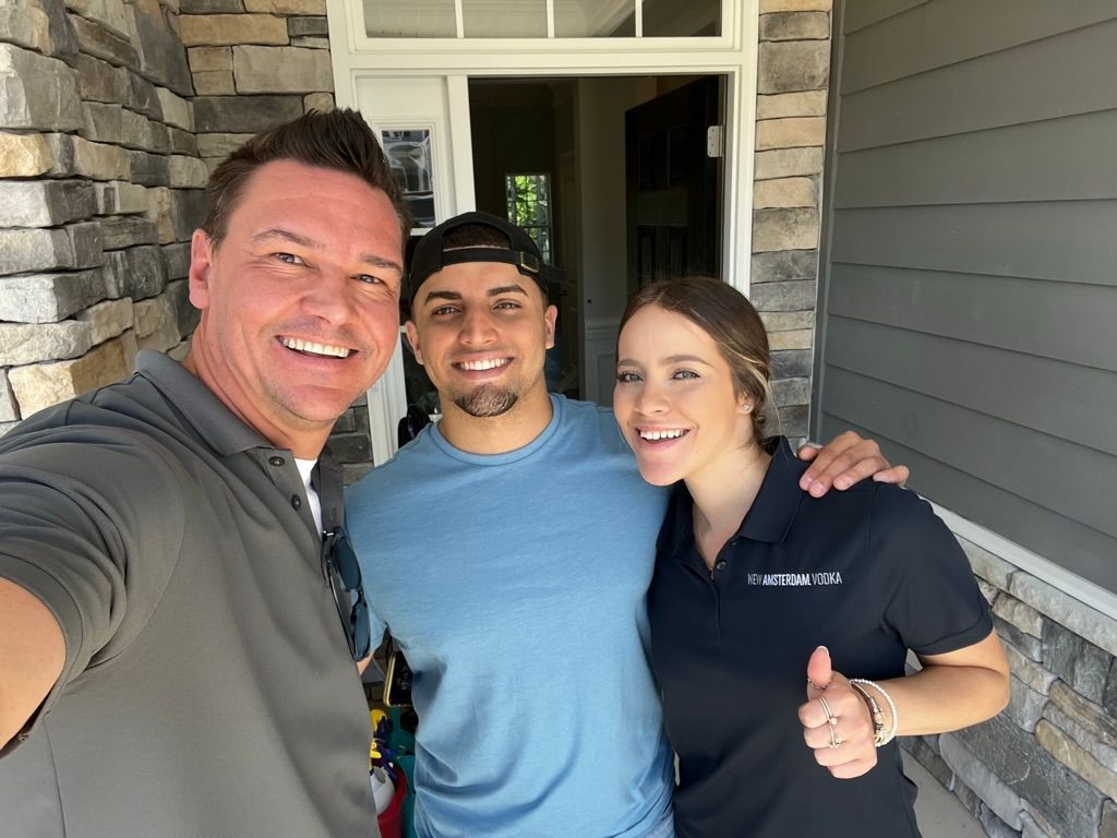 Three smiling people posing on a porch; one gives a thumbs-up.