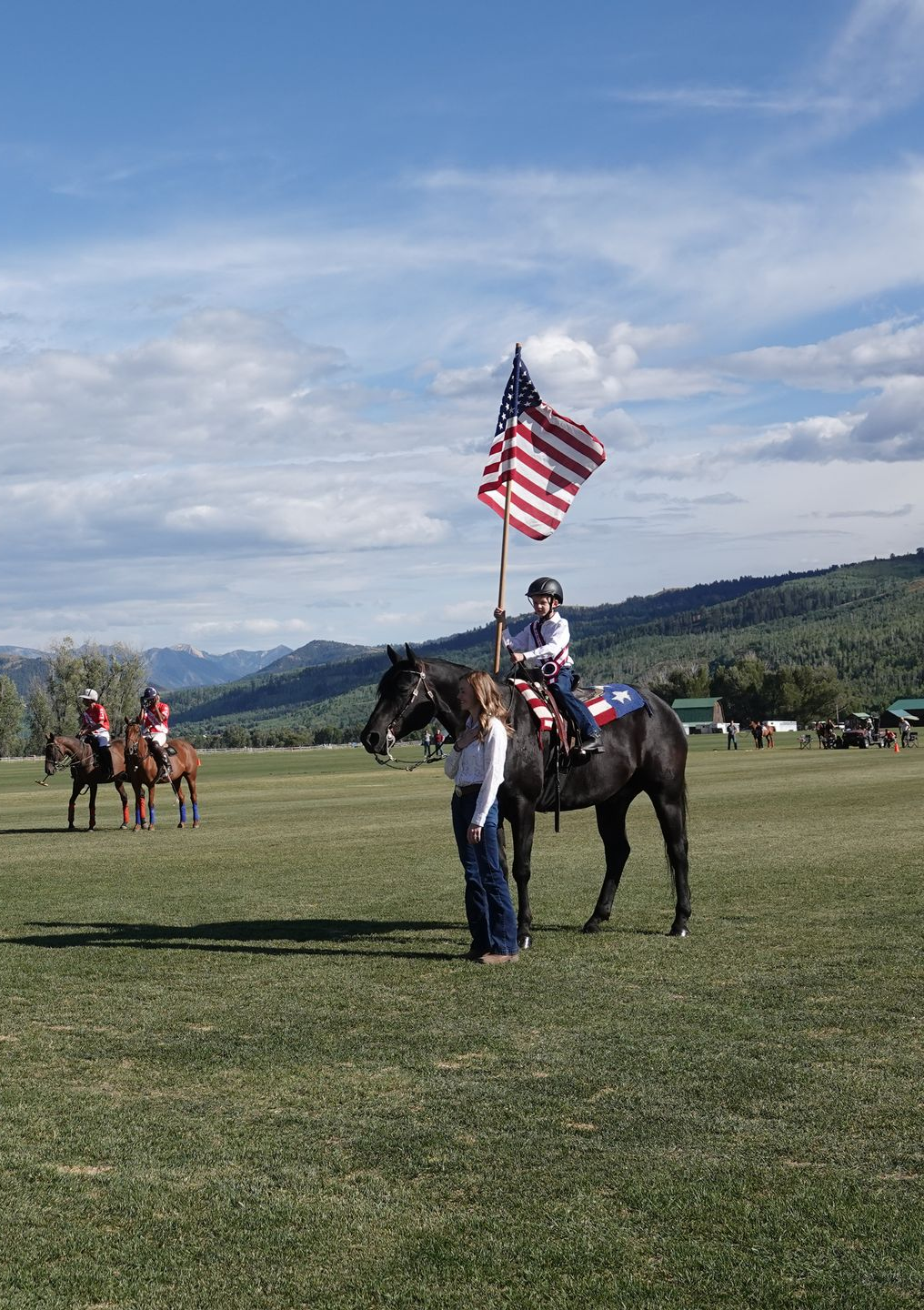 Child on horseback holding American flag, supported by a person. Horses and riders in background, grassy field, mountains.