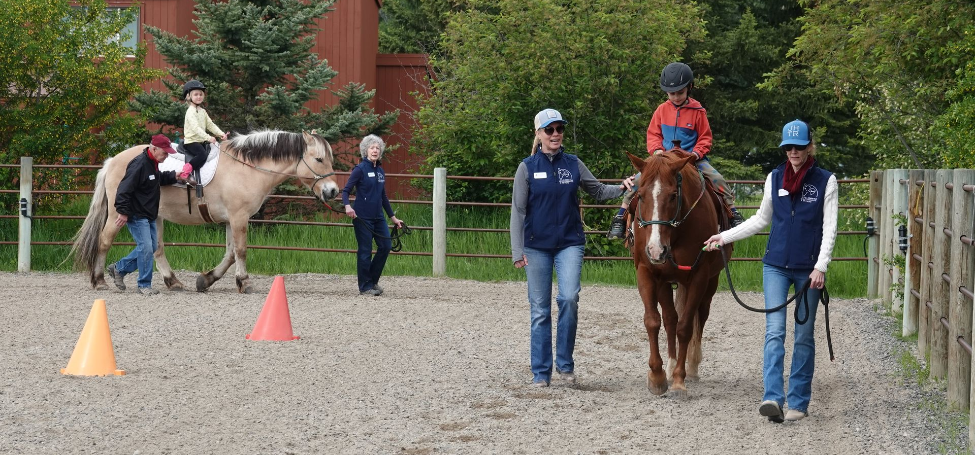 Instructors guide two children riding horses in an outdoor riding arena with orange safety cones on the ground.