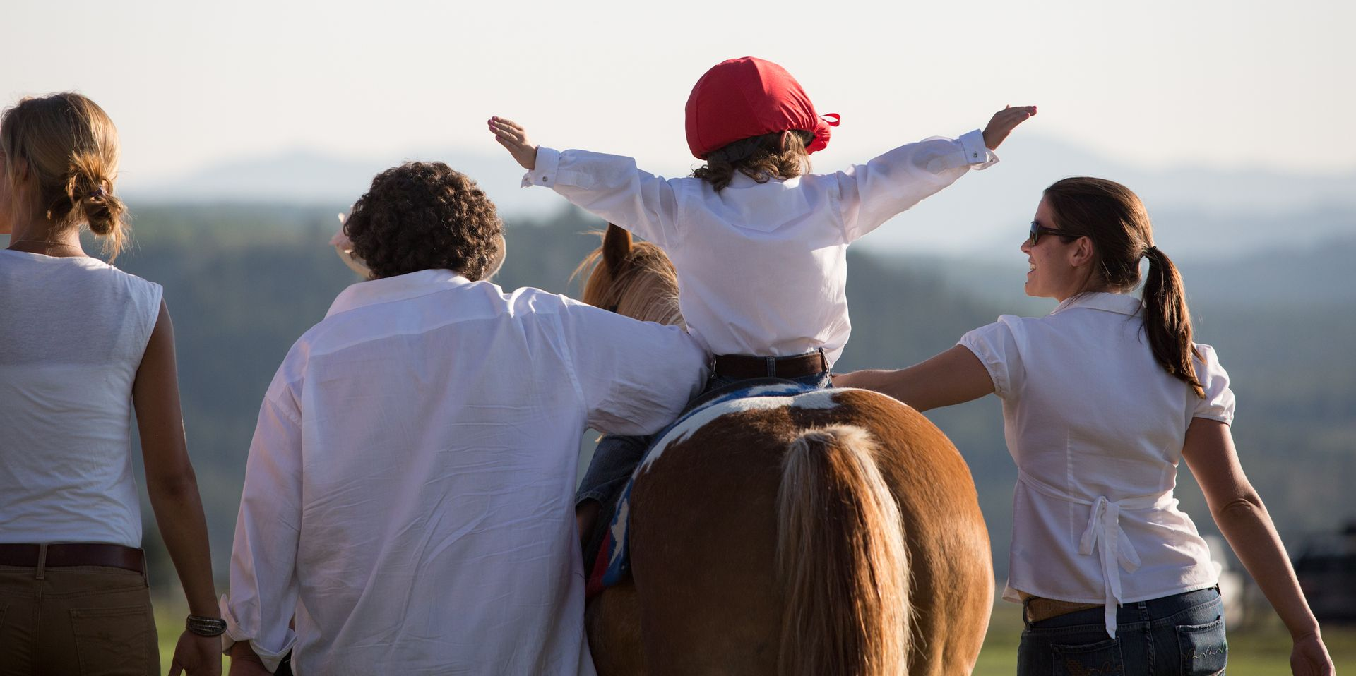 A child in a red helmet with arms outstretched rides a brown horse, flanked by two adults walking on a sunny day.