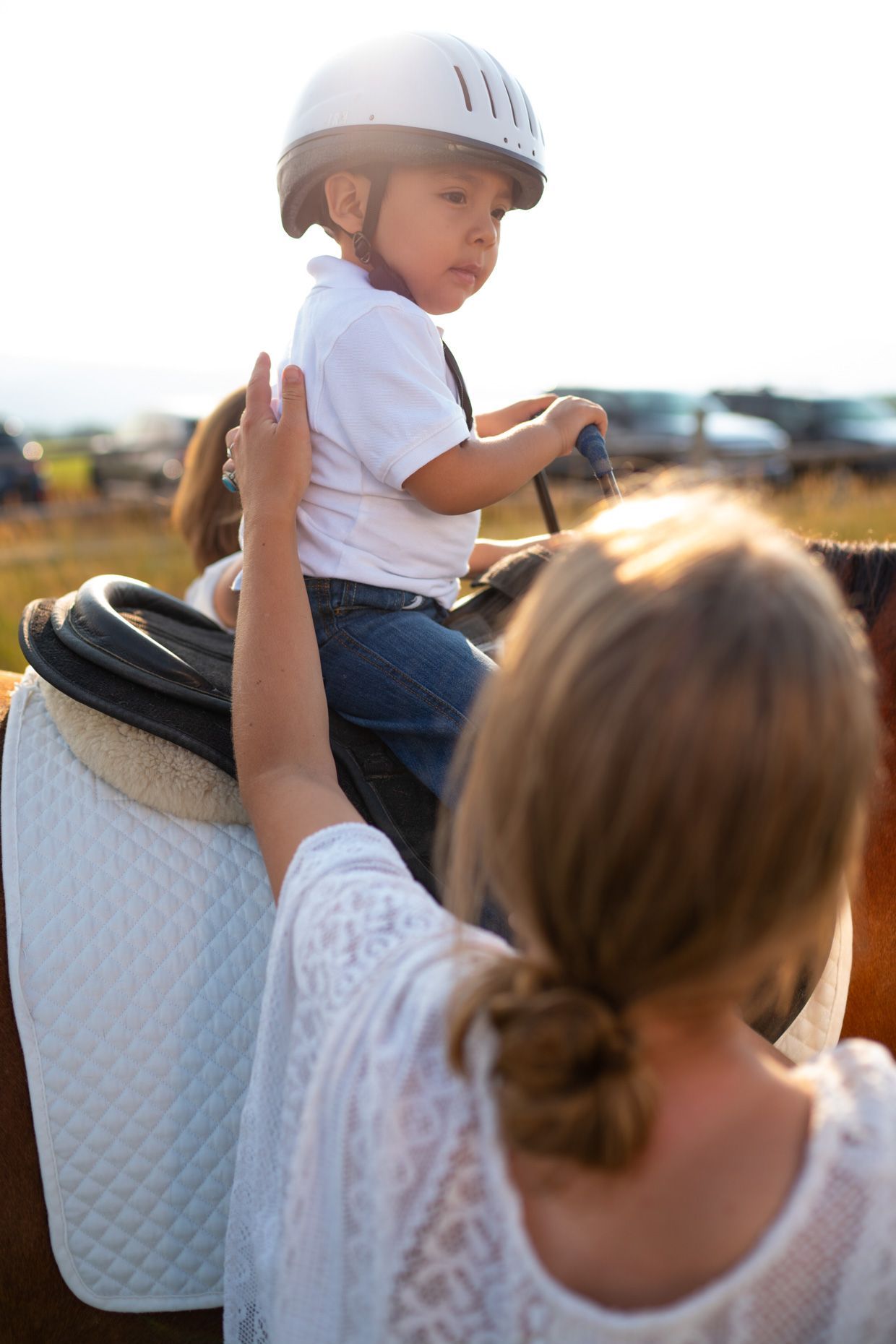 A person in a white top stands behind a child wearing a helmet, who is sitting on a saddled horse in an outdoor setting.