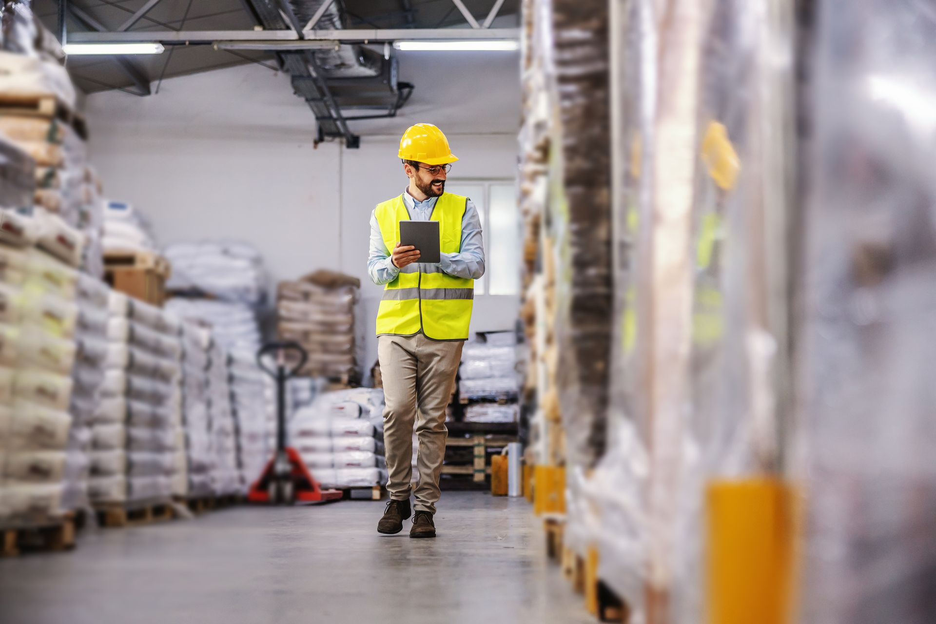 Warehouse worker in a yellow vest and hard hat, using a tablet, walking down an aisle with inventory. Warehouse worker in a yellow vest and hard hat, using a tablet, walking down an aisle with inventory.