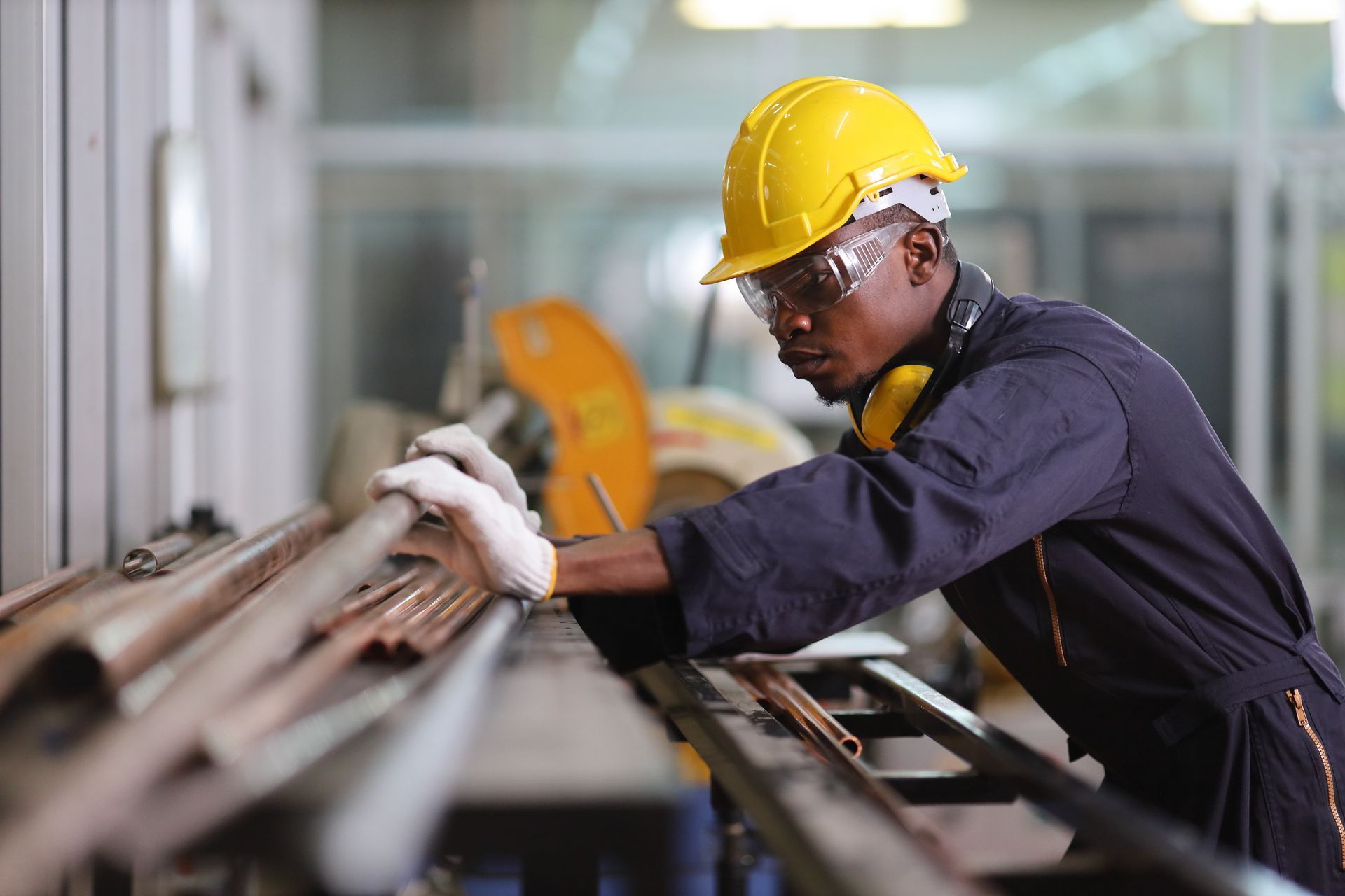 Factory worker in yellow hard hat, safety glasses, and gloves working with metal.