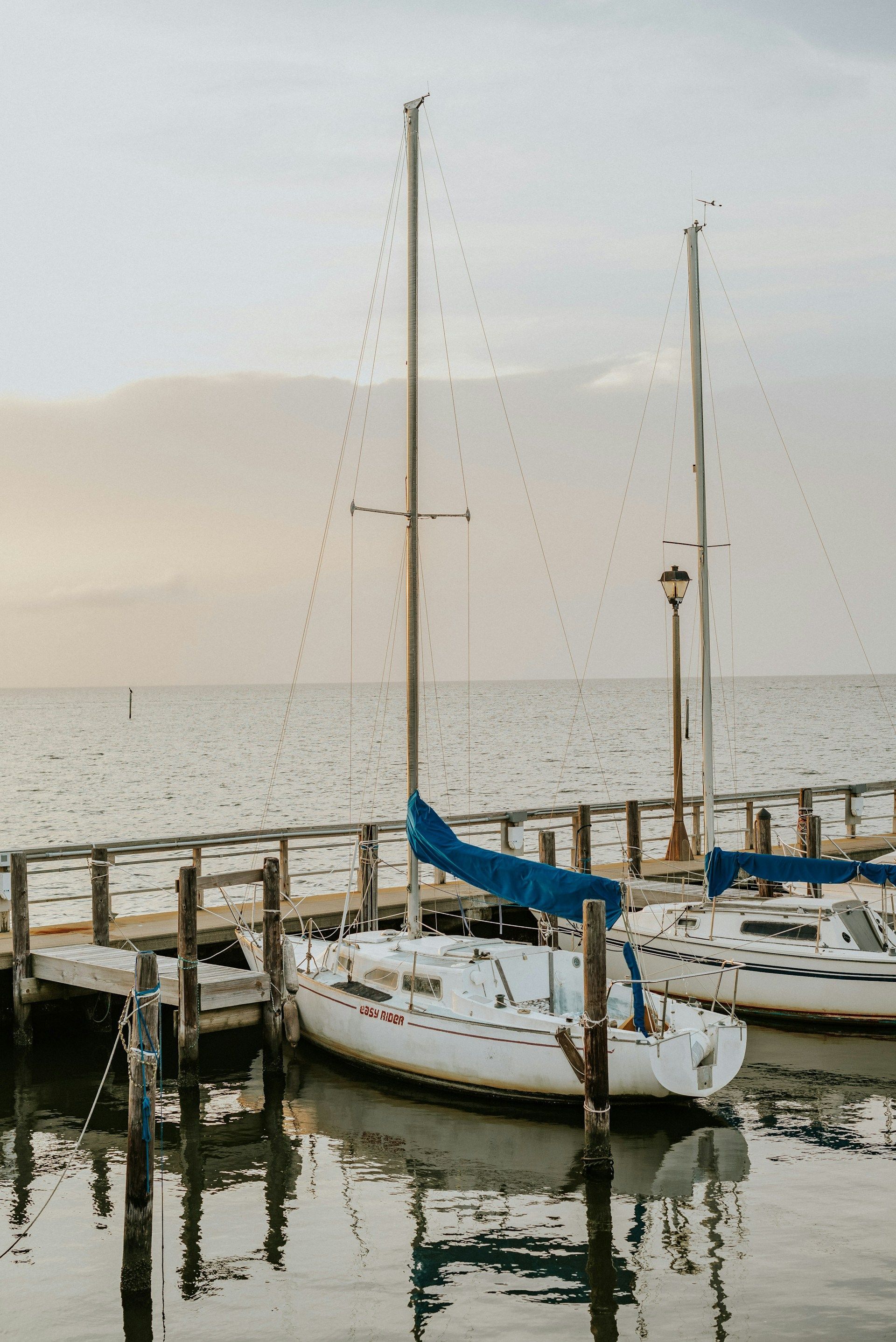 Sailboats docked at a wooden pier on calm water; sky is pale yellow.