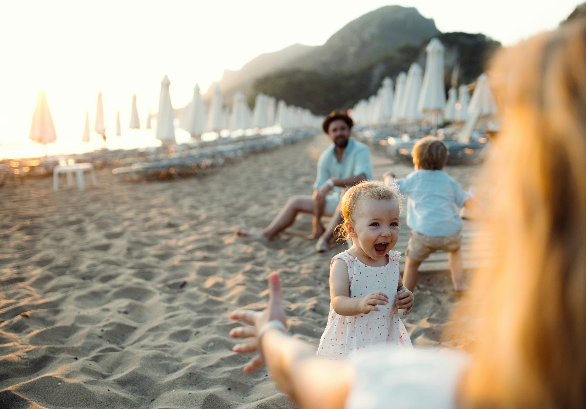 Woman with outstretched arms, toddler runs towards her, beach setting, man and child in background.