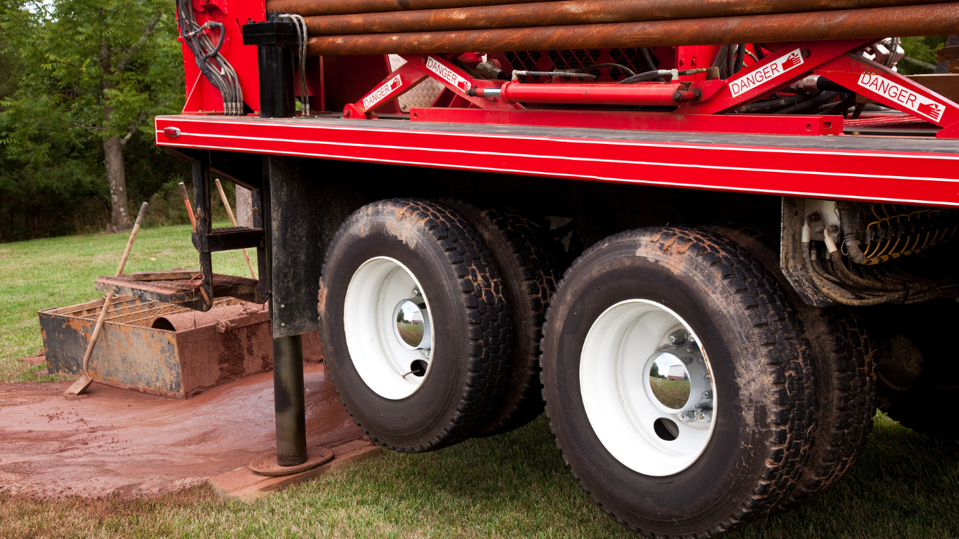 Red drilling rig with white-rimmed tires on grass, drilling into the ground.