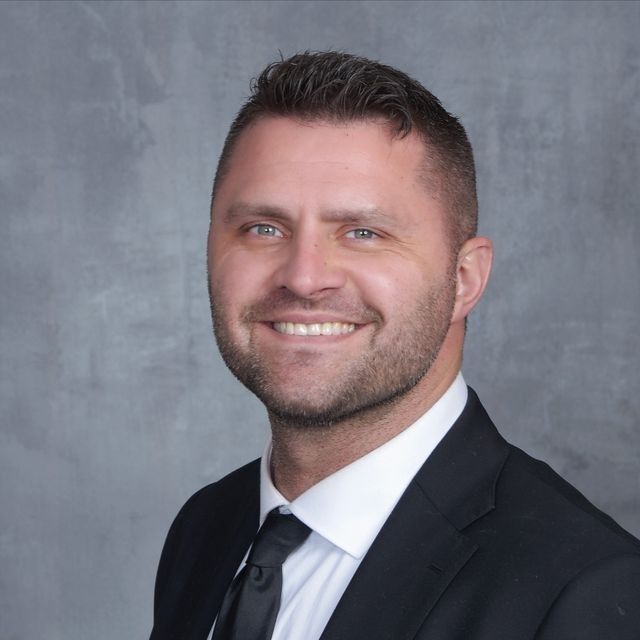Portrait of a smiling man in a black suit and tie against a gray background