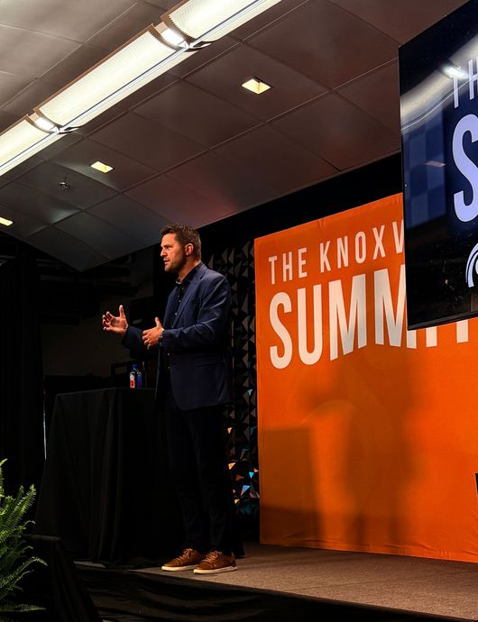 Man speaking on stage with a microphone beside a screen reading “Leadership Beyond the Scoreboard”