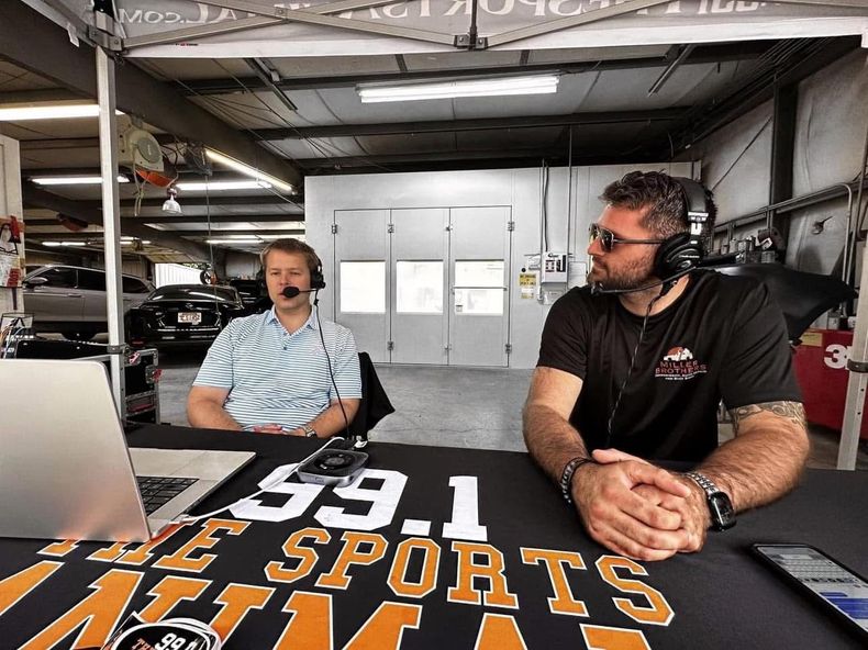 Two men at a radio studio table with microphones and a “Sports” banner in an industrial garage setting
