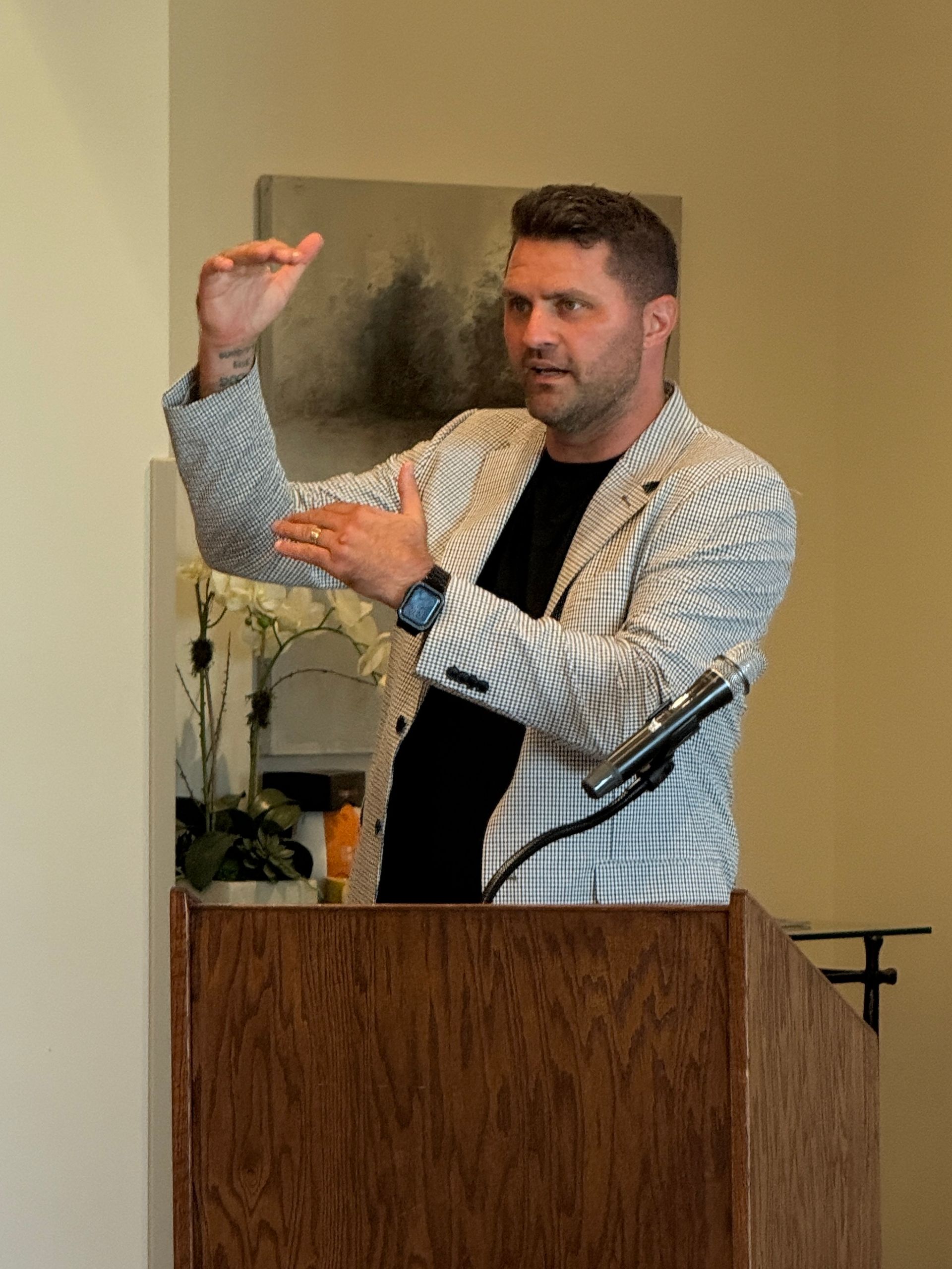 Man in black speaking on stage, holding a microphone and gesturing with one hand